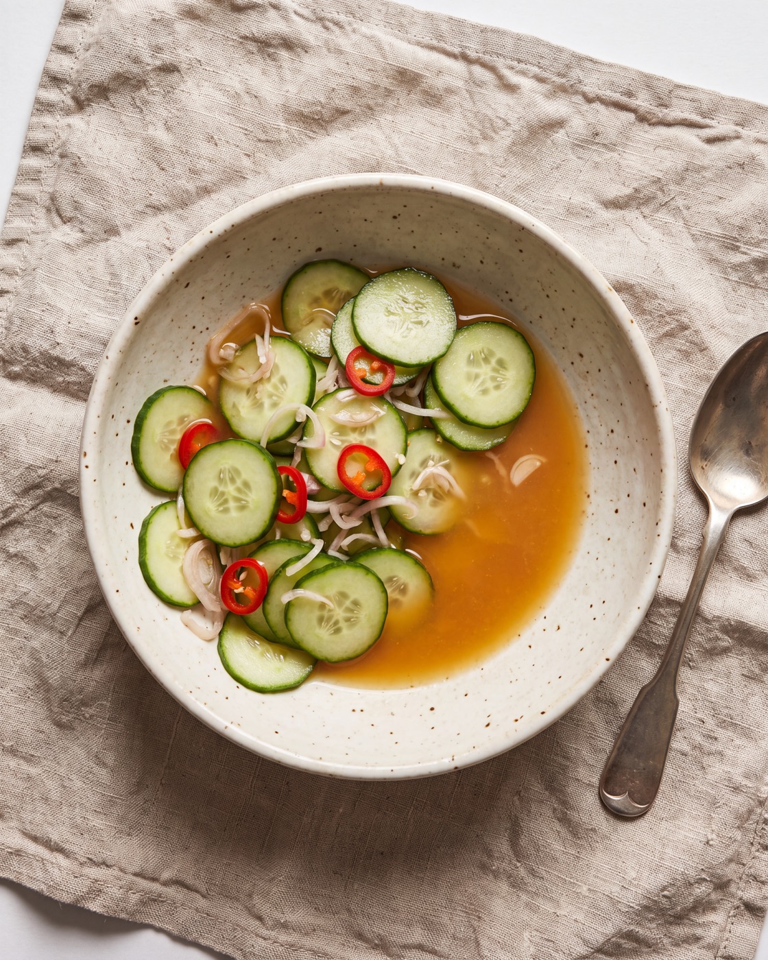 Cucumber, shallot and chilli tossed loosely in a cloudy amber kink pickling liquid, in a speckled bowl with a spoon beside it