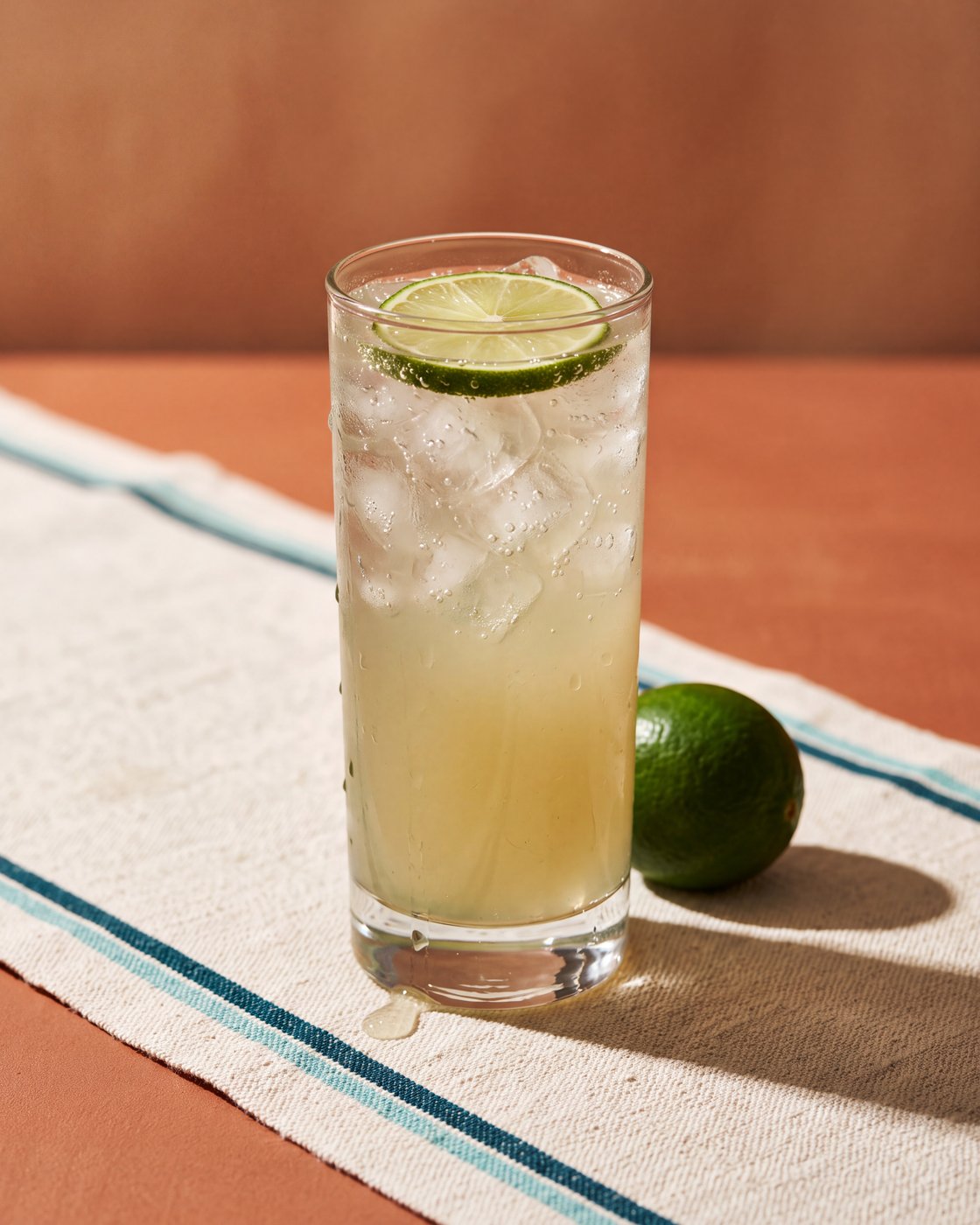 kink Ginger Lime Sparkling filled with ice, condensation on the glass, a lime wheel on top and a whole lime beside it on a woven cotton runner