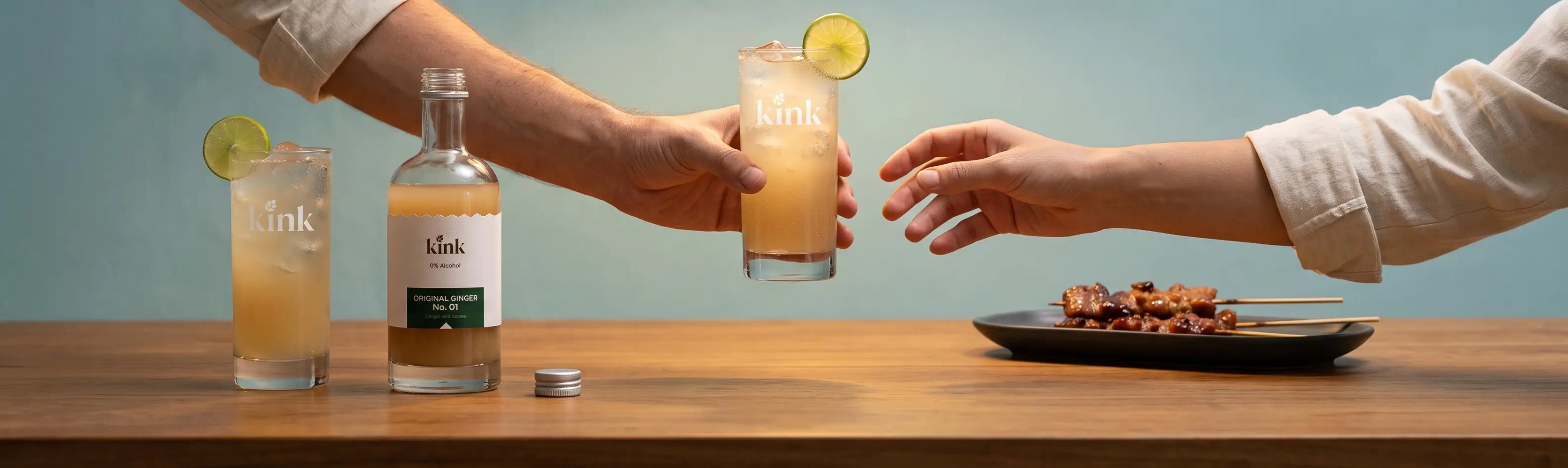 Hands passing a glass of kink ginger soda across a sunlit table, beside the bottle, more drinks and sharing plates